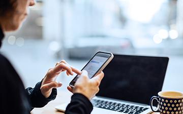 Woman wearing a black shirt, sitting behind a laptop while navigating on her smartphone